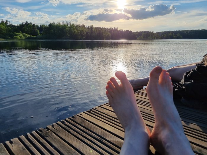 Två personers fötter på en träbrygga med en sjö och skog i solnedgång i bakgrunden.