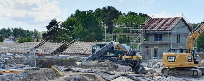 Byggplats med ett nästan färdigt hus under ett sadeltak och flera omonterade tak på marken.