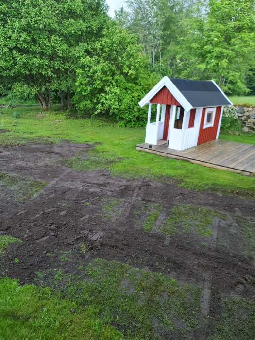 Ojämn gräsmatta under renovering med synlig jord och grävspår, litet rött lekhus och gröna träd i bakgrunden.