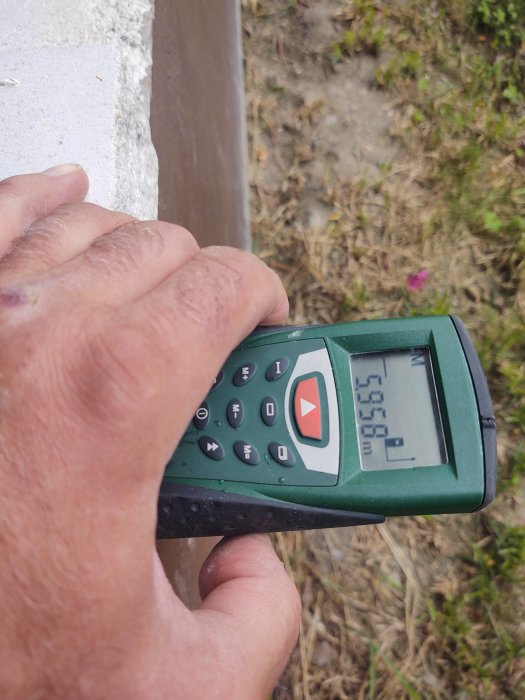 Hand håller en lasermätare med mätresultatet 5.98 m, med fasadtegel och gräs i bakgrunden.
