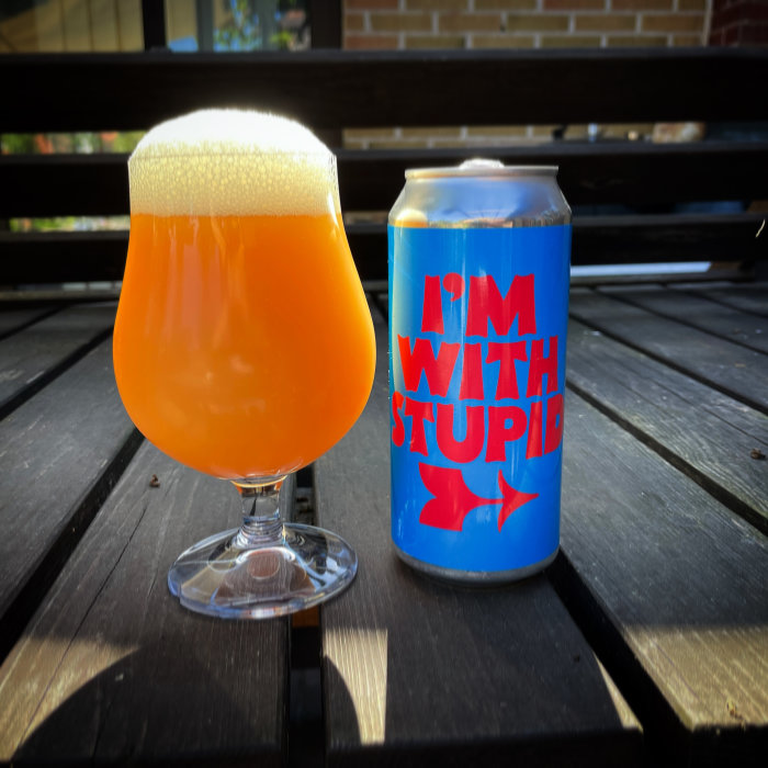 Orange dryck i glas vid sidan av burk med texten "I'M WITH STUPID" på en träbänk.