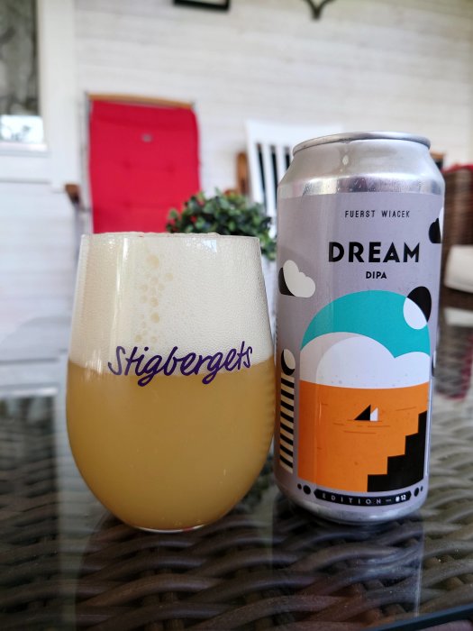 Ölglas med skum från Stigbergets Bryggeri och en burk DIPA på ett rottingbord med en röd dörr i bakgrunden.