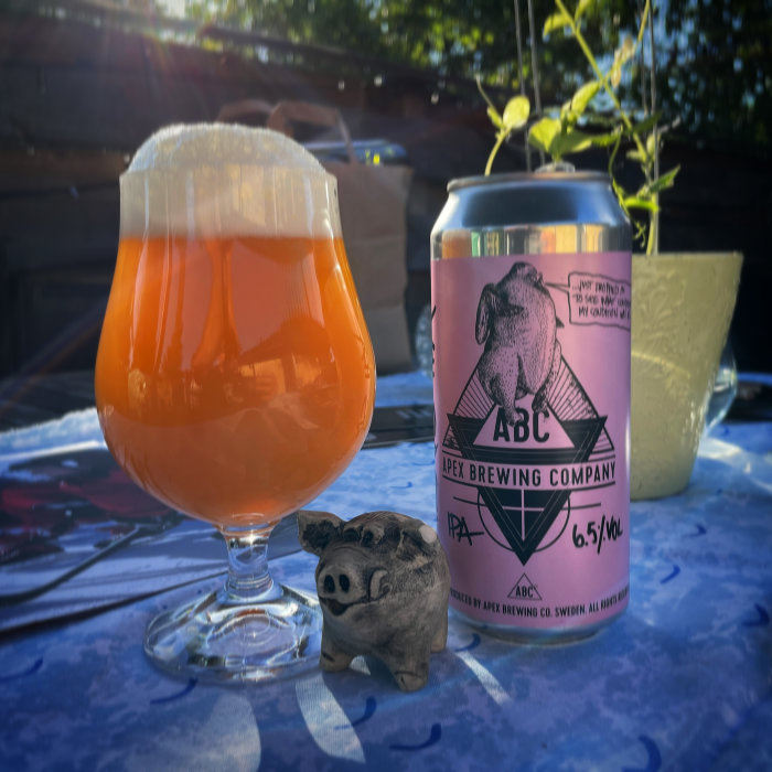 Ölglas fyllt med orangefärgad dryck och en ölburk från Apex Brewing Company på ett bord utomhus.