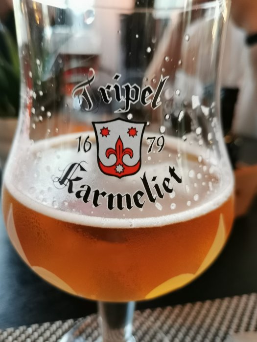 Ett glas Tripel Karmeliet öl delvis drucket på ett bord.