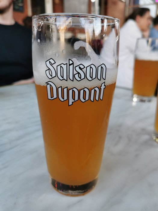 Halvdrucken öl i ett glas märkt "Saison Dupont" på ett bord, med människor i bakgrunden.
