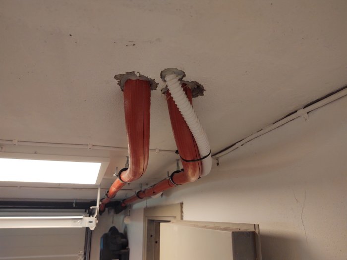 Isolerade rör och en vit kondensvattenslang monterade längs ett tak i ett pannrum.