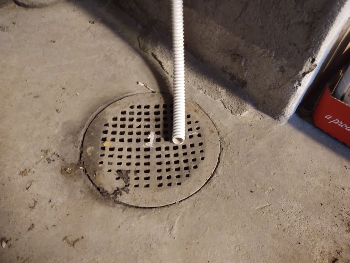 Vit kondensvattenslang ansluten till en brunnslock på ett betonggolv, vilket illustrerar dräneringssystemet.