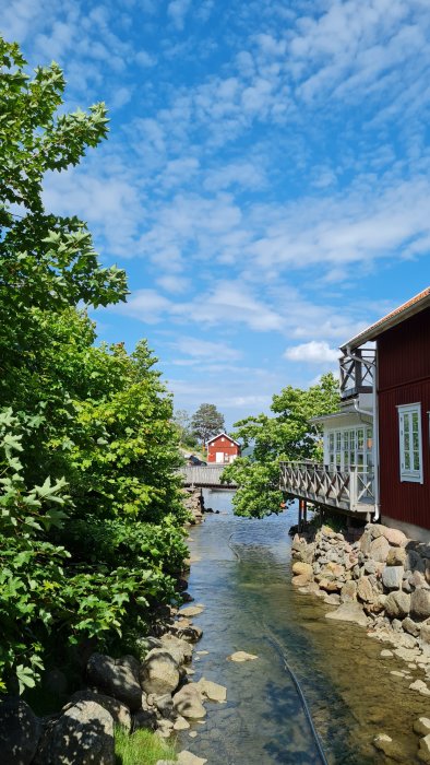 Idyllisk vy av en träbrygga och röda stugor vid en klar bäck på västkusten under en blå himmel med vita moln.