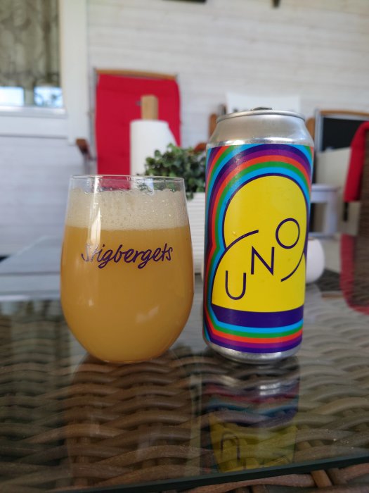 Ölglas märkt med Stigbergets fyllt med grumlig öl och en burk med Omnipollo-design på ett glasbord.