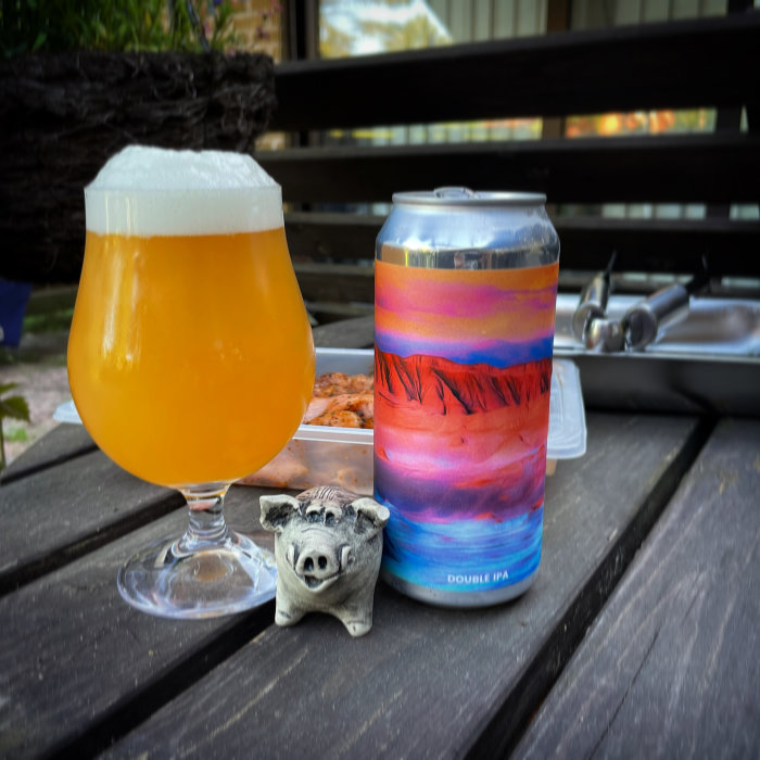 Ett glas med skummande öl bredvid en färgstark burk Double IPA på ett träbord utomhus.