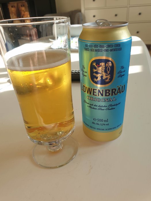 Ett glas med guldfärgad öl bredvid en blå och gul Löwenbräu ölburk på ett bord.