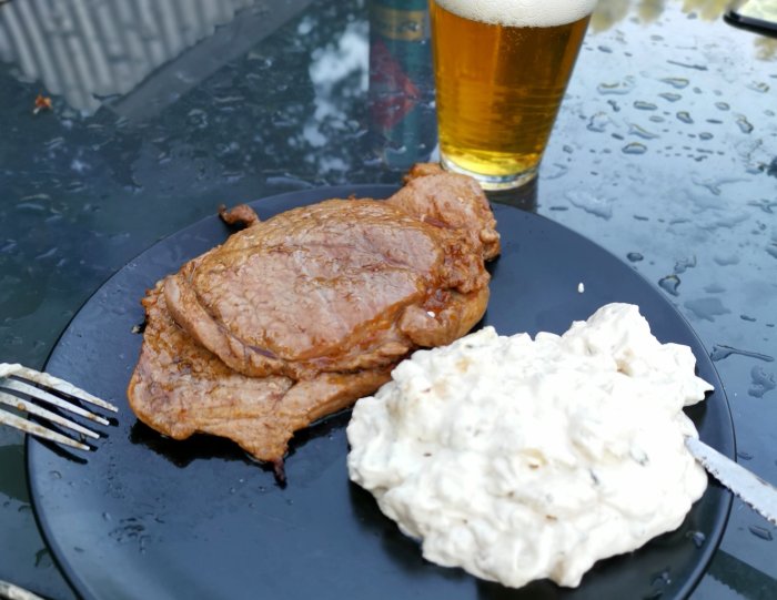 Grillad biff och potatissallad på svart tallrik med ölglas i bakgrunden.