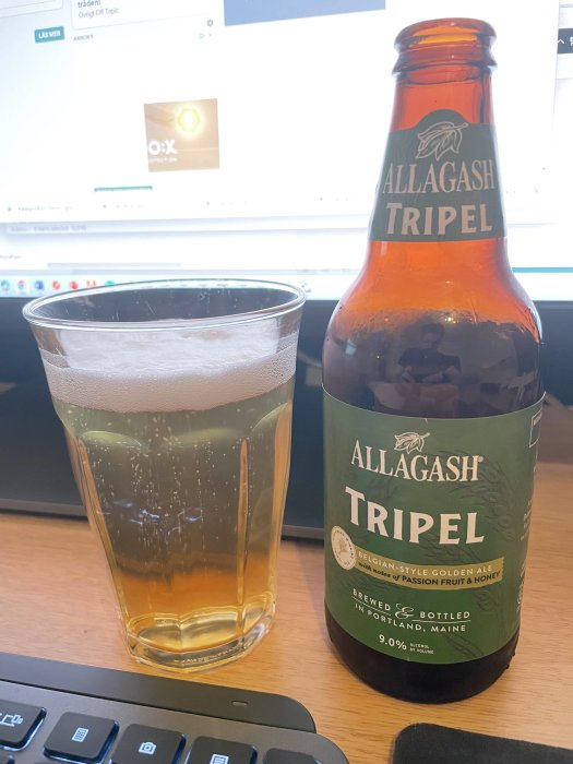 Ölglas med ljus öl bredvid en Allagash Tripel flaska på ett skrivbord med datorskärm i bakgrunden.