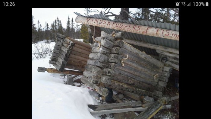 Sönderslaget vindskjul av trä med bråte och snö runt, texten "VINDSKYDD FÖRSÄLJN" syns.