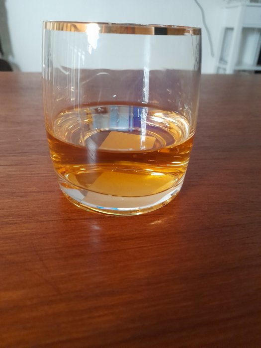 Whiskyglas halvfullt med gulbrun dryck på trämöblerad yta.