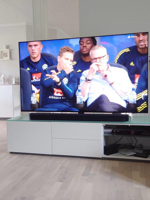 TV-skärm som visar en fotbollsmatch med spelare och tränare på avbytarbänken.