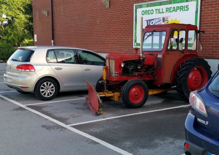 Röd veterantraktor parkerad bredvid en silverfärgad bil på en parkeringsplats.