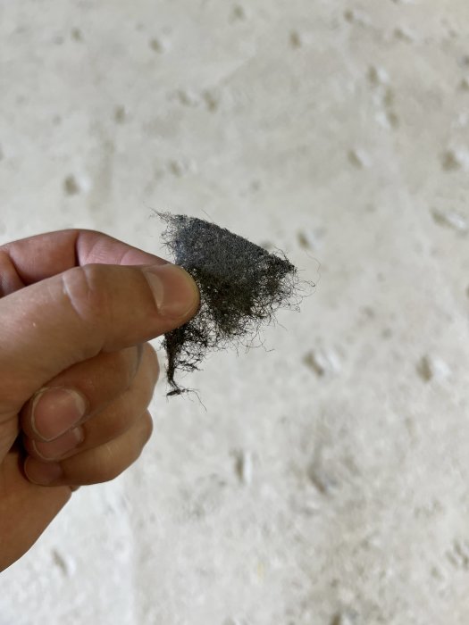 Hand håller ett stycke svart fibermaterial mot en suddig bakgrund av ett betonggolv.