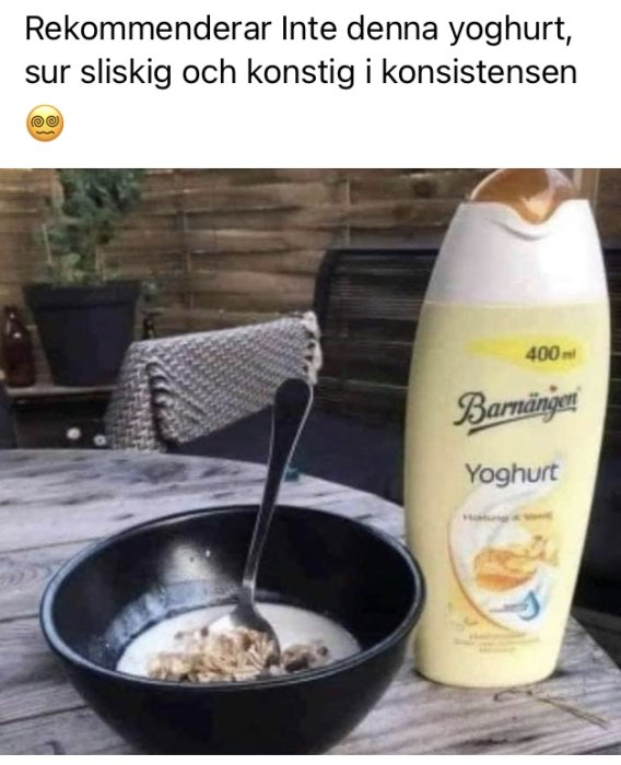 Svart skål med müsli och en sked fast i tjock yoghurt bredvid en flaska av "Bamajongt" yoghurt på ett bord.