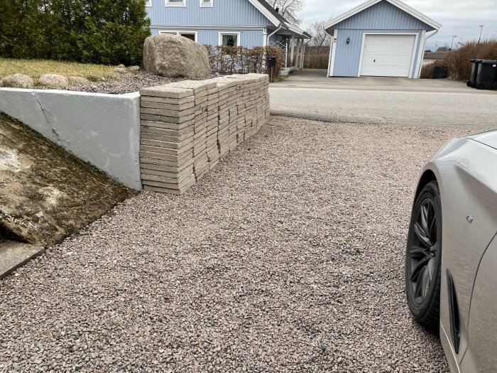 Stapel med stora grå trottoarplattor intill uppfart med grusbotten framför ett hus och garage.