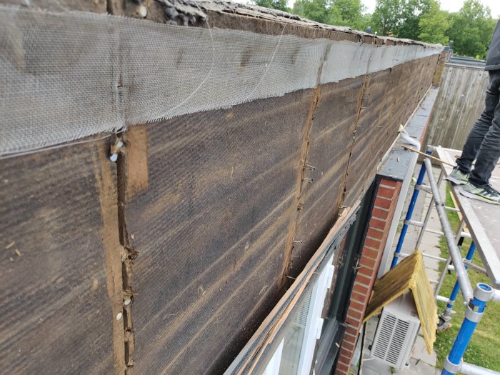 Närbild på en husvägg där gammal träpanel är borttagen, visar asfaboard utan luftspalt och delar av byggnadsställning.