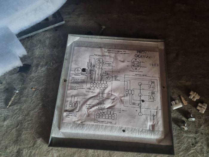 Nedfläckat elschema monterat i genomskinlig plastförpackning på ett dammigt underlag med lösa plastdelar runtomkring.
