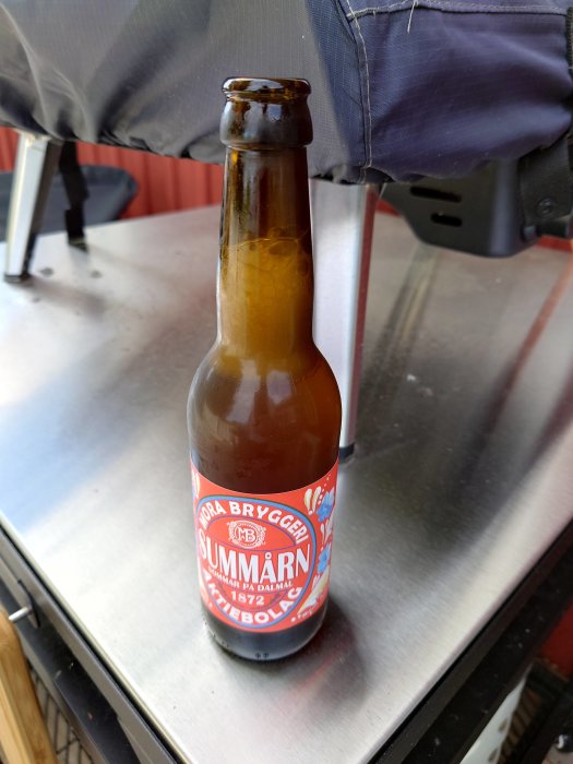 En ölflaska från Mora Bryggeri märkt "Sommarn" står på en metallisk yta med en grill i bakgrunden.
