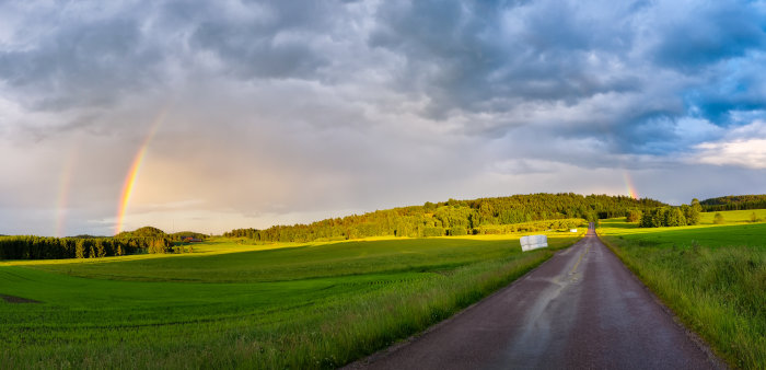 Landsväg som sträcker sig mot en dubbel regnbåge över gröna fält och skog.