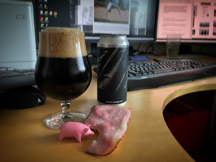 Ölglas med skum på skrivbord, burk med texten "BLACK DOUBLE IPA", datormus och tangentbord, plastgris på skinkbit.