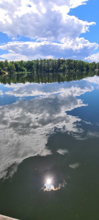 Speglingsbild av moln och träd i det stilla vattnet i Magelungen med solreflektion, tagen mitt på dagen.