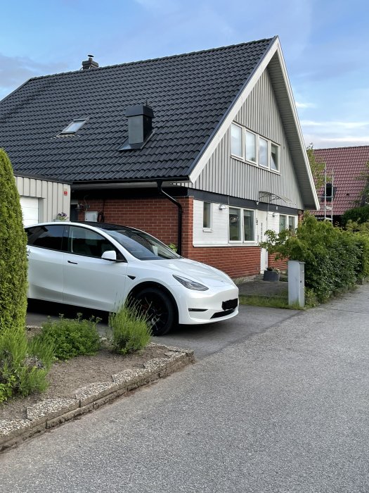 En vit elbil parkerad vid en garagebyggnad bredvid huset med synlig laddbox monterad på garagets sidovägg.