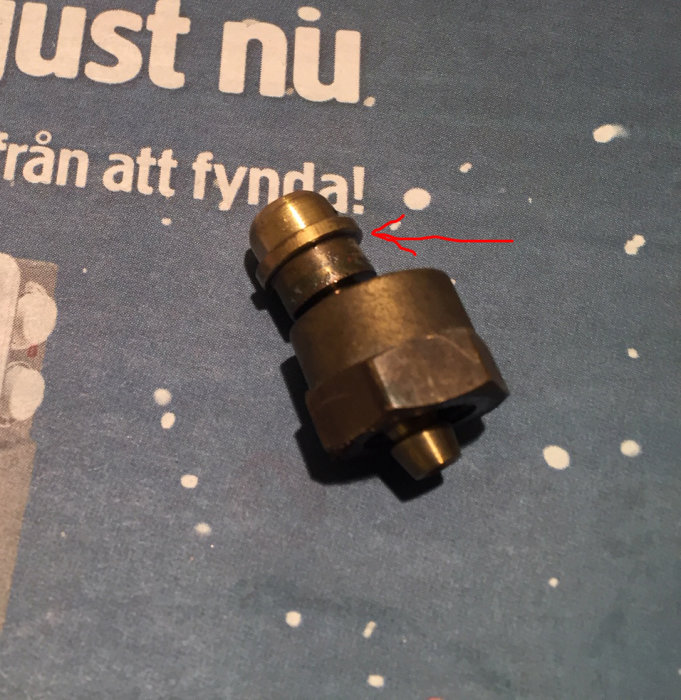 Gaskabelnippel demonterad från en husvagn, placerad på ett tryckt mönstrat underlag, med en pil som pekar på skarven.