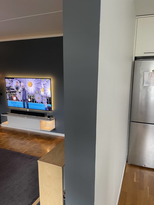 Hörn av en bärande grå vägg i ett hem, nära ett vardagsrum med TV och en vit köksinredning.