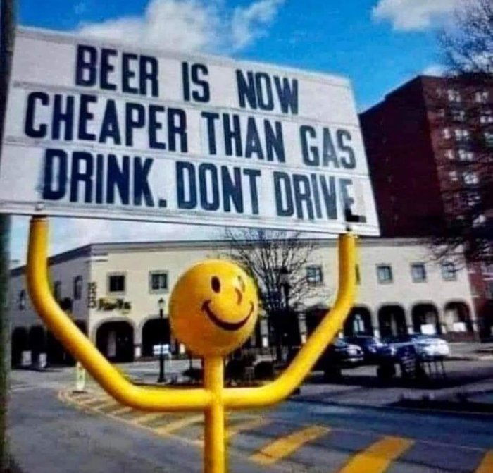 Skylt med texten "Beer is now cheaper than gas. Drink. Don't drive" ovanför ett gatuföremål med leende ansikte.