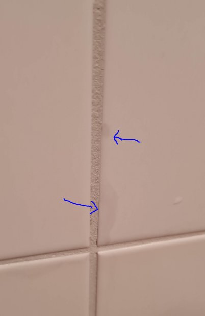 Närbild på vit kakelfog med hårfina sprickor markerade med blå pilar.