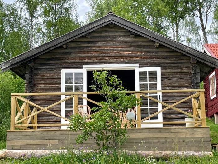 Renoverad timmerstuga med nybyggd träveranda och öppet fönster omgiven av grönska.