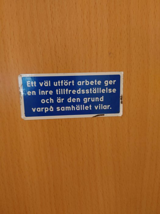 Blå skylt med texten "Ett väl utfört arbete ger en inre tillfredsställelse och är den grund varpå samhället vilar" på träyta.