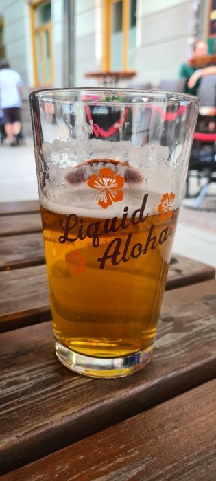 Halvfullt ölglas märkt "Liquid Aloha" på ett träbord utomhus, med suddiga personer i bakgrunden.