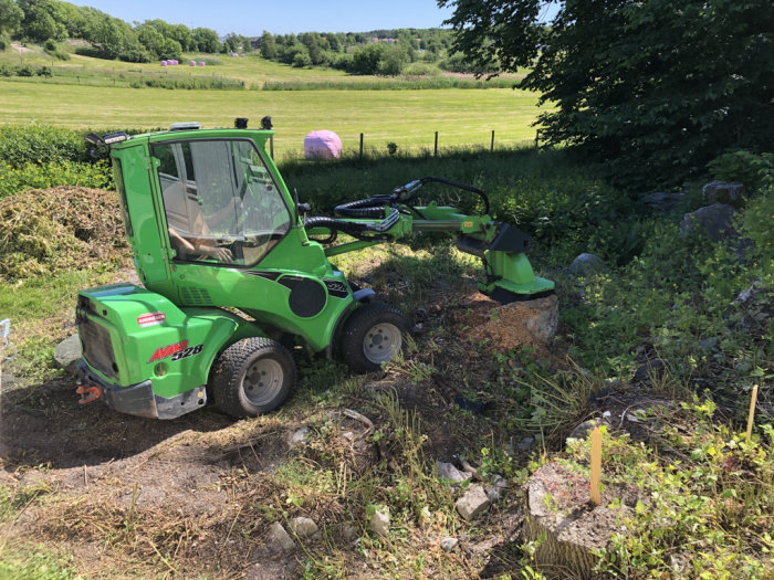 Grön Avant styrbar minilastare med stubbfräs i arbete på en solig äng.