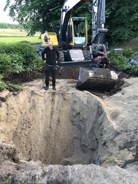 Person framför grävmaskin vid ett stort hål i jorden på en byggplats för en altan.