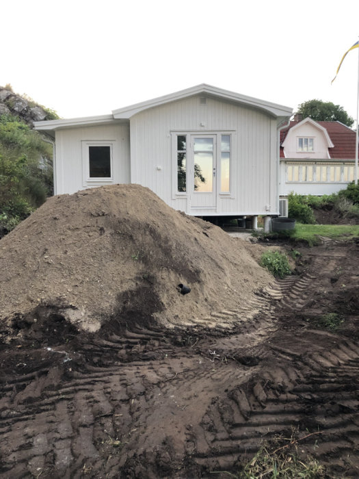 Hög med grävd jord framför vit villa under altanbygge, med bandspår från grävmaskin i förgrunden.