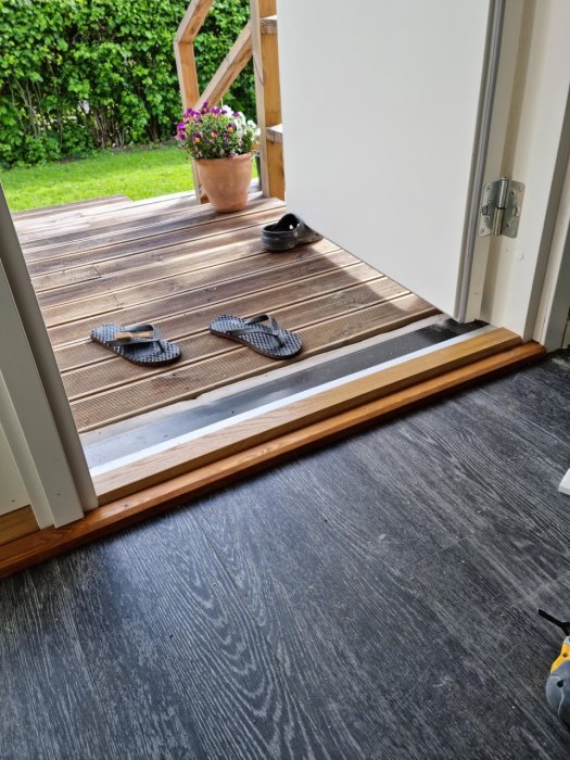 Nyinstallerad trätröskel vid ytterdörr övergång till mörkt golv med sandaler på trädäck och krukväxt utanför.