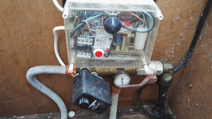 En vattenpumps kontrollpanel med diverse kablar, en tryckströmbrytare och tryckmätare, tecken på korrosion och åldrade komponenter.