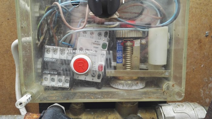 Transparent elektrisk kopplingsbox med sladdar och en röd startknapp ovanpå en vattenpump, som indikerar ett problem beskrivet i inlägget.