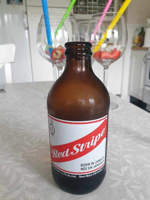 Brun flaska Red Stripe-öl med suddiga vinglas och sugrör i bakgrunden på ett bord.