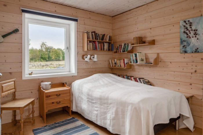 Sovrum med liggande träpanel på väggarna, inbyggda bokhyllor, trämöbler och ett fönster med utsikt.