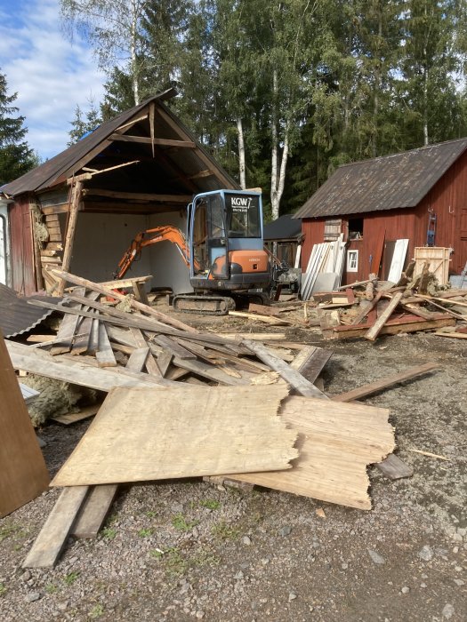 Renoveringsarbete med grävmaskin vid en delvis riven byggnad och träspill på marken.