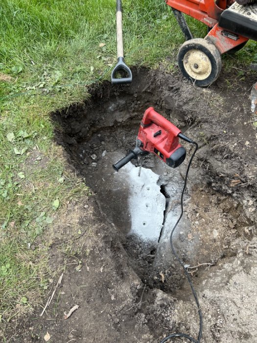 Grävd grop i trädgård med redskap, en röd borrmaskin, och en teknik för att bryta sten med grillkol och kopplade luftslangar.