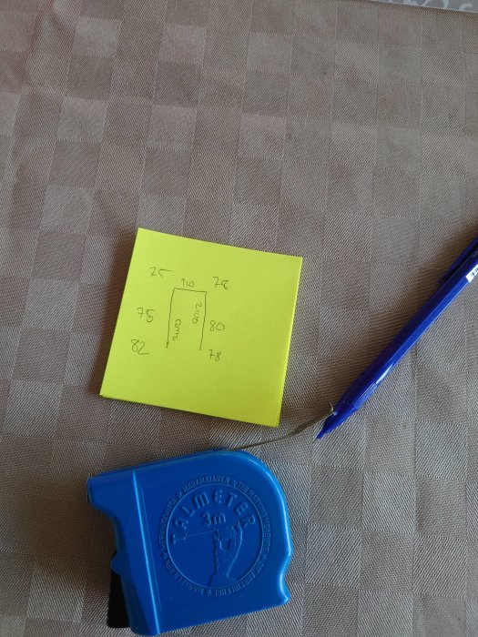 Måttanteckningar på en gul post-it lapp bredvid en blå måttbandshållare och en blå penna på ett rutigt underlag.