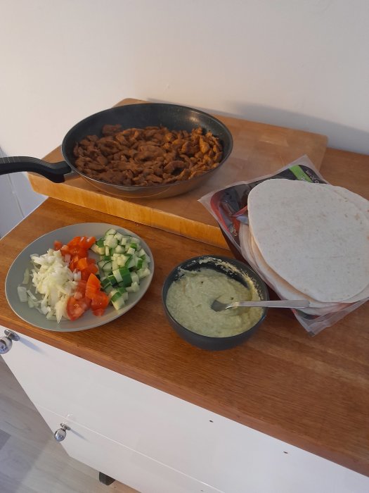 Ingredienser för att göra tacokebab: stekt kött i en panna, tortillabröd och hackade grönsaker på en skärbräda.